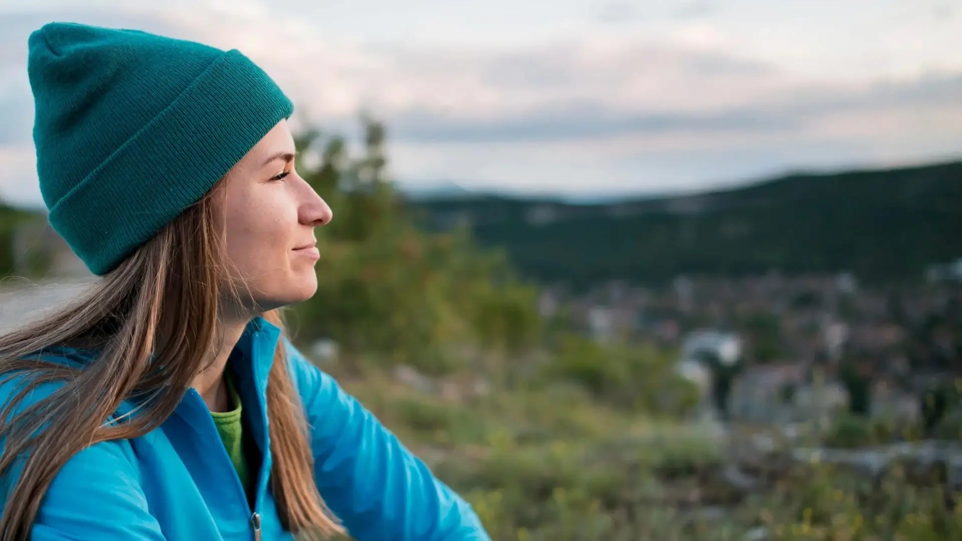 A woman in a teal beanie and blue jacket gazing thoughtfully outdoors with a blurred natural background.