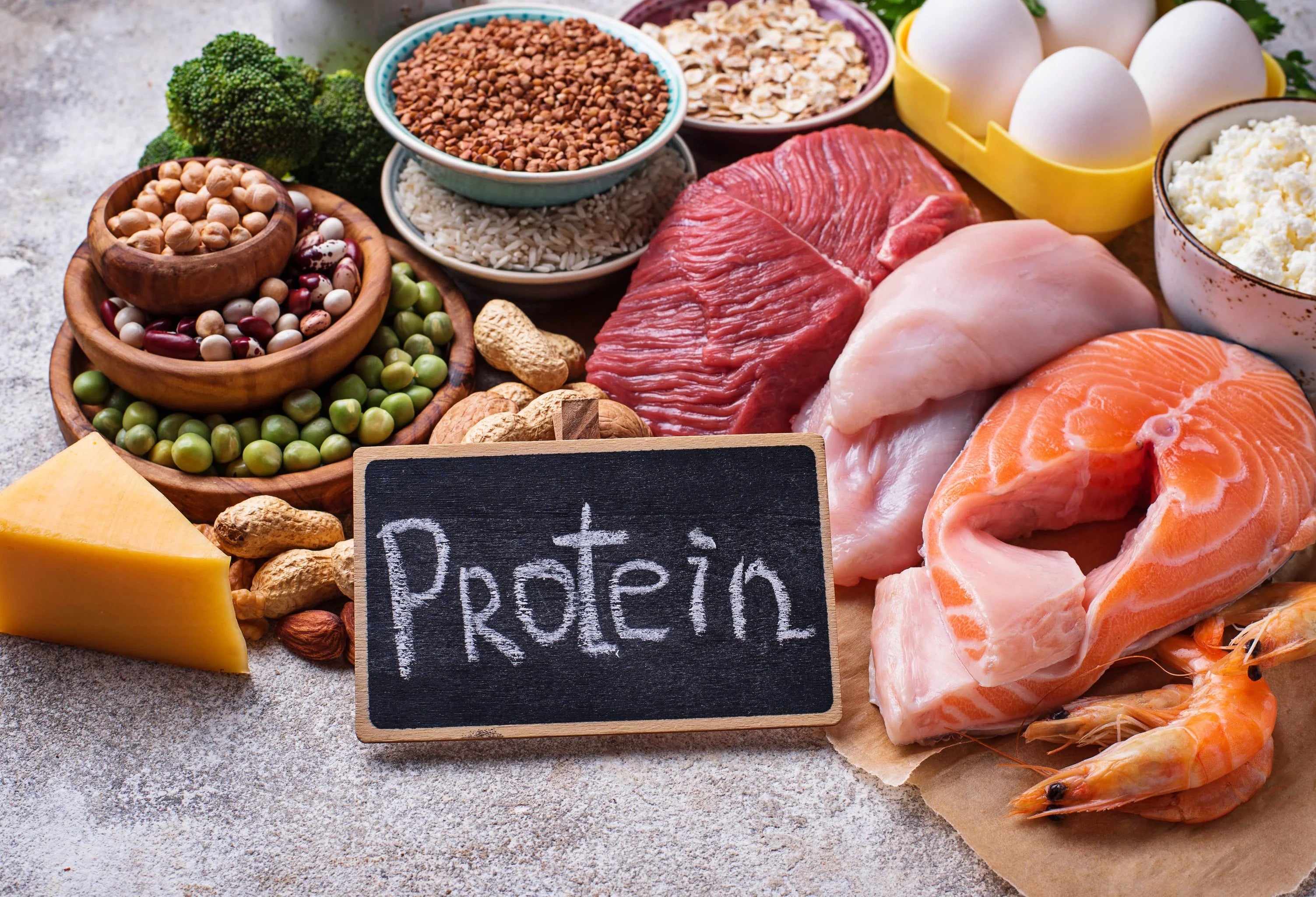 A sign with 'Protein' written on it with chalk and various protein rich foods soch as salmon, shrimp, chicken breast, beef, eggs, cheese, various grains