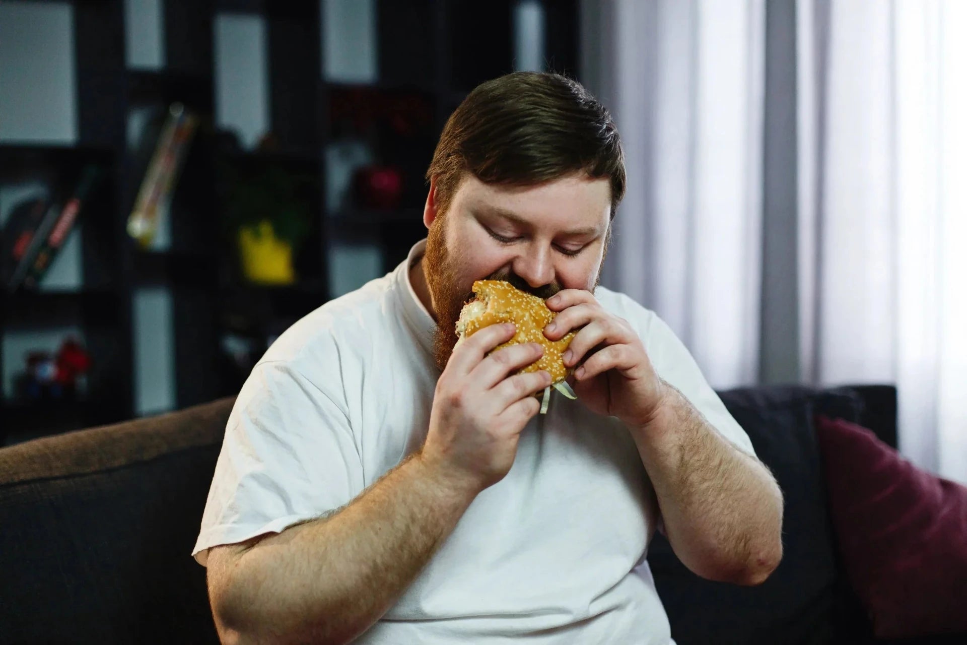 Bearded man eating hamburger while sitting on a sofa.